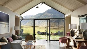 A spacious living room with a high ceiling, large windows offering a mountain view, a cozy seating area with a sofa and chairs, and a wooden coffee table. - Eleven Deplar Farm - All inclusive (Skagafjörour)