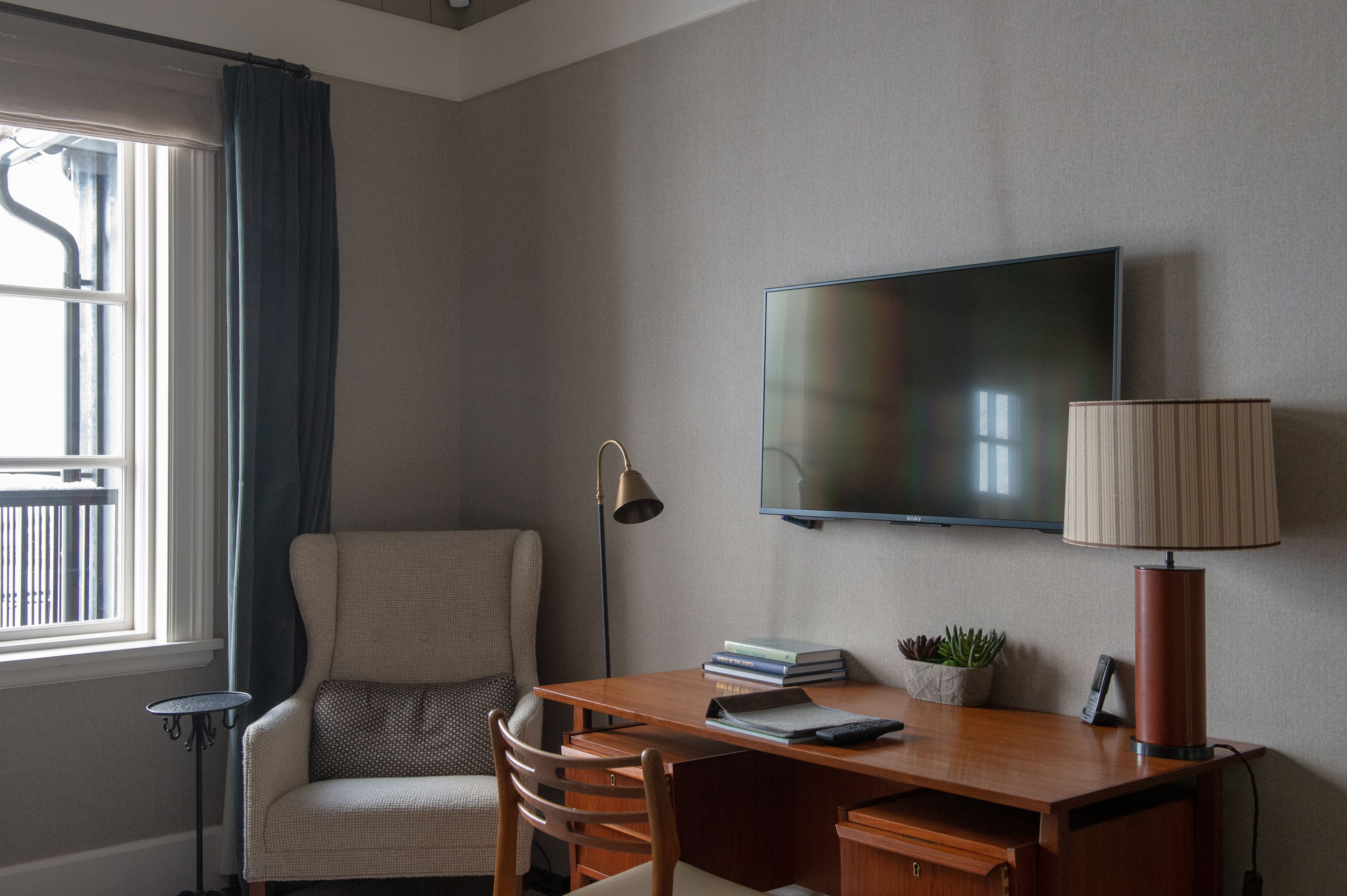 A hotel room with a large flat-screen TV mounted on the wall, a wooden desk with a lamp, a chair, and a small table.