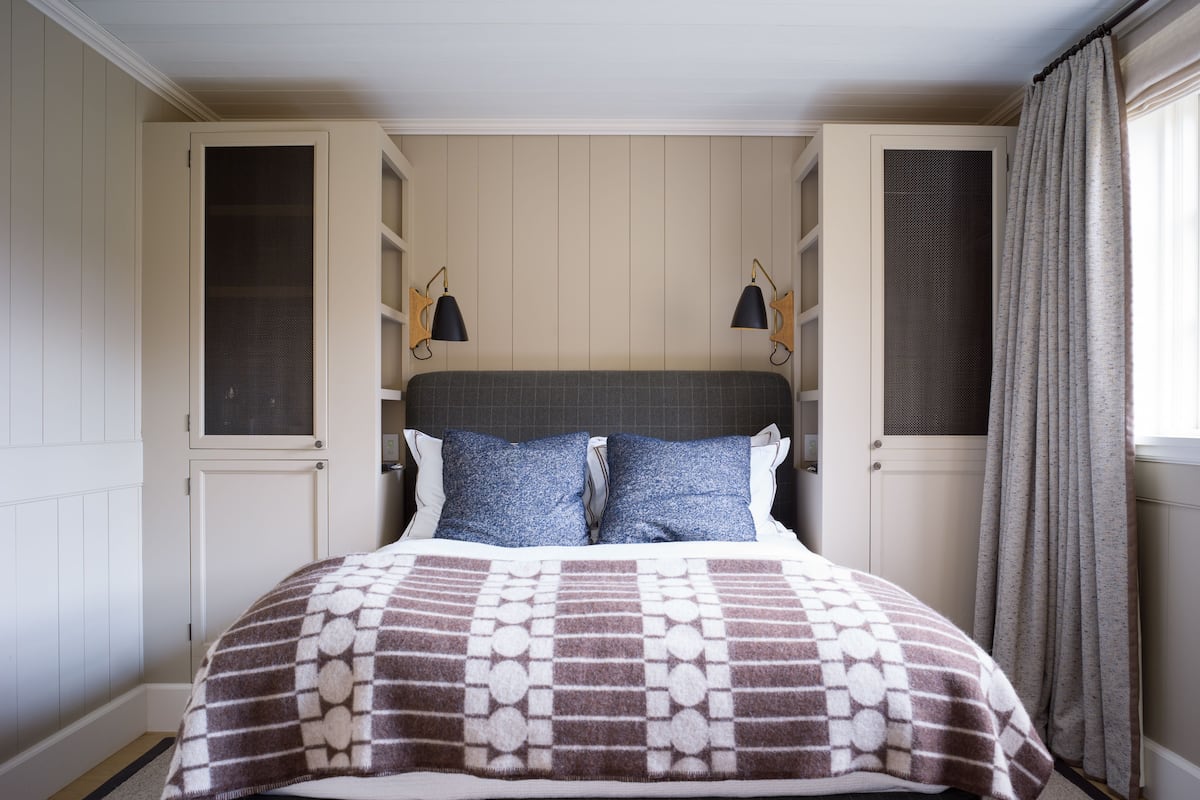 Un lit bien fait, recouvert d’une couvre-lit à motifs, deux lampes fixées au mur et une fenêtre avec des rideaux.