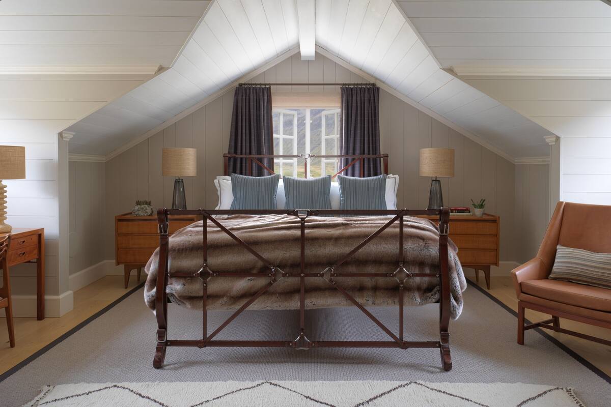 Une chambre avec un plafond en pente, un lit avec un cadre en bois, deux tables de chevet avec des lampes, une chaise et une fenêtre avec des rideaux.