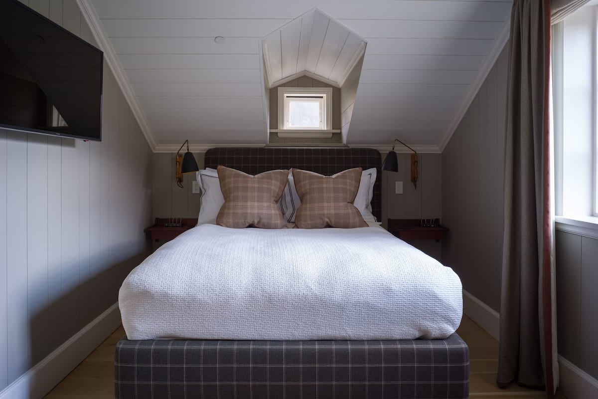 Une chambre à coucher comprenant un lit, une table de chevet, une lampe, une fenêtre avec des rideaux et un plafond en pente doté d’un puits de lumière.