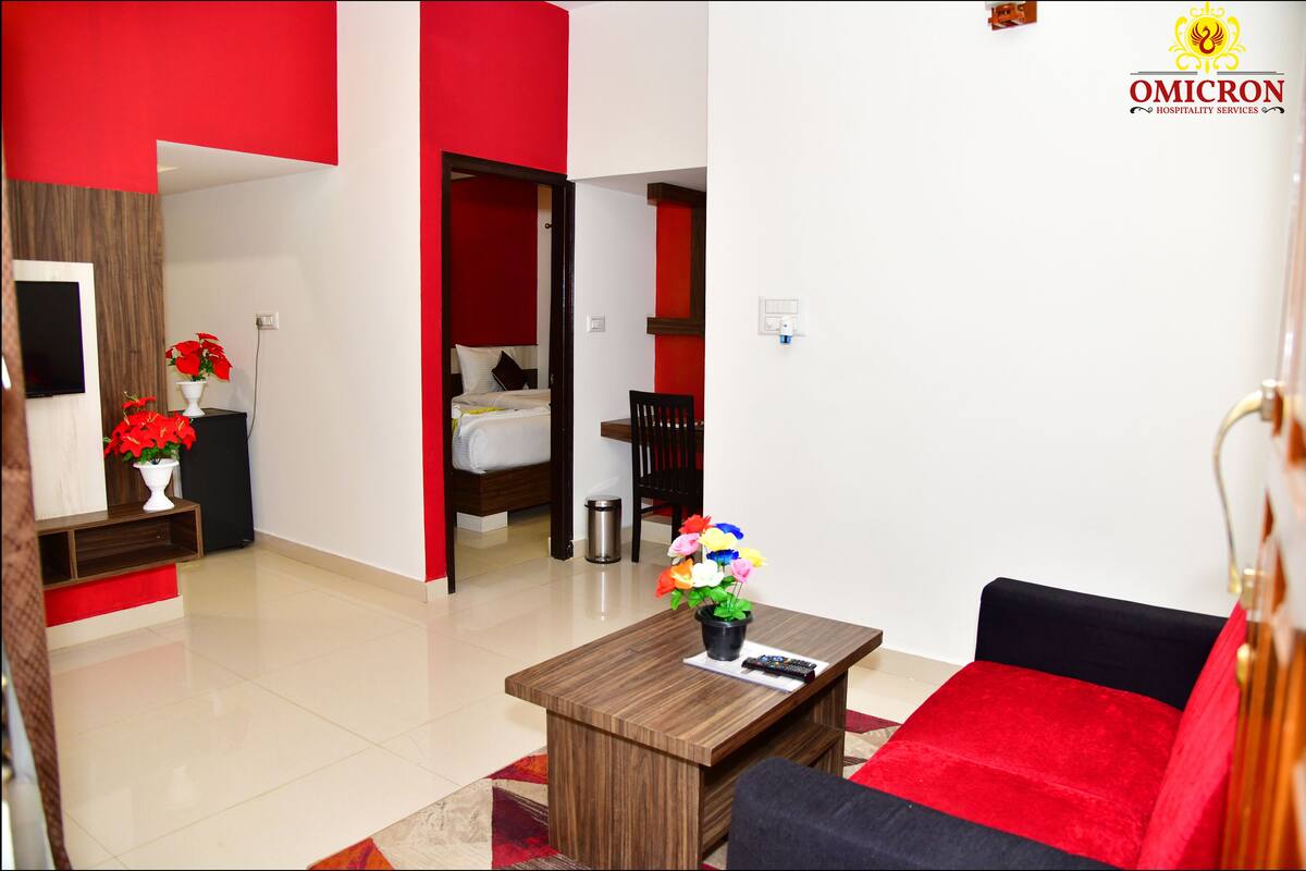 A modern hotel room with a red and white color scheme, a wooden coffee table with a vase of flowers, a black sofa, and a bed with a red accent.