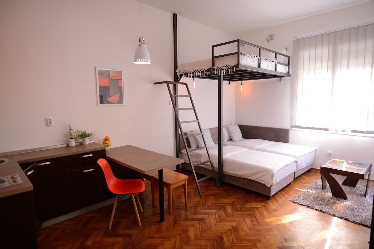 A compact room with a bunk bed, a desk with a chair, a small table, and a window with curtains.