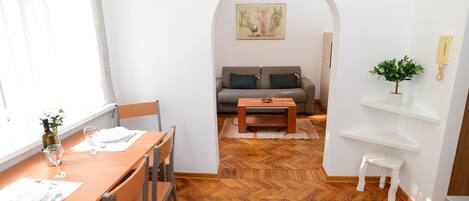 A dining area with a wooden table and chairs, a sofa in the background, and a small white stool.