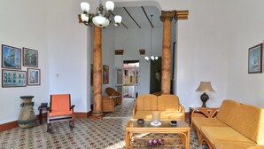 A spacious living room with a patterned floor, a sofa, a chair, a coffee table, and a lamp. There are framed pictures on the walls and a large window in the background. - Hostal Belvédère (Havana)