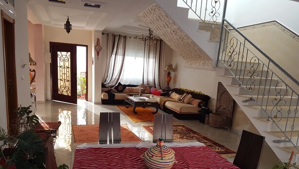 Un salón espacioso con una escalera, una zona de comedor con mantel a rayas rojas, y un sofá con cojines.