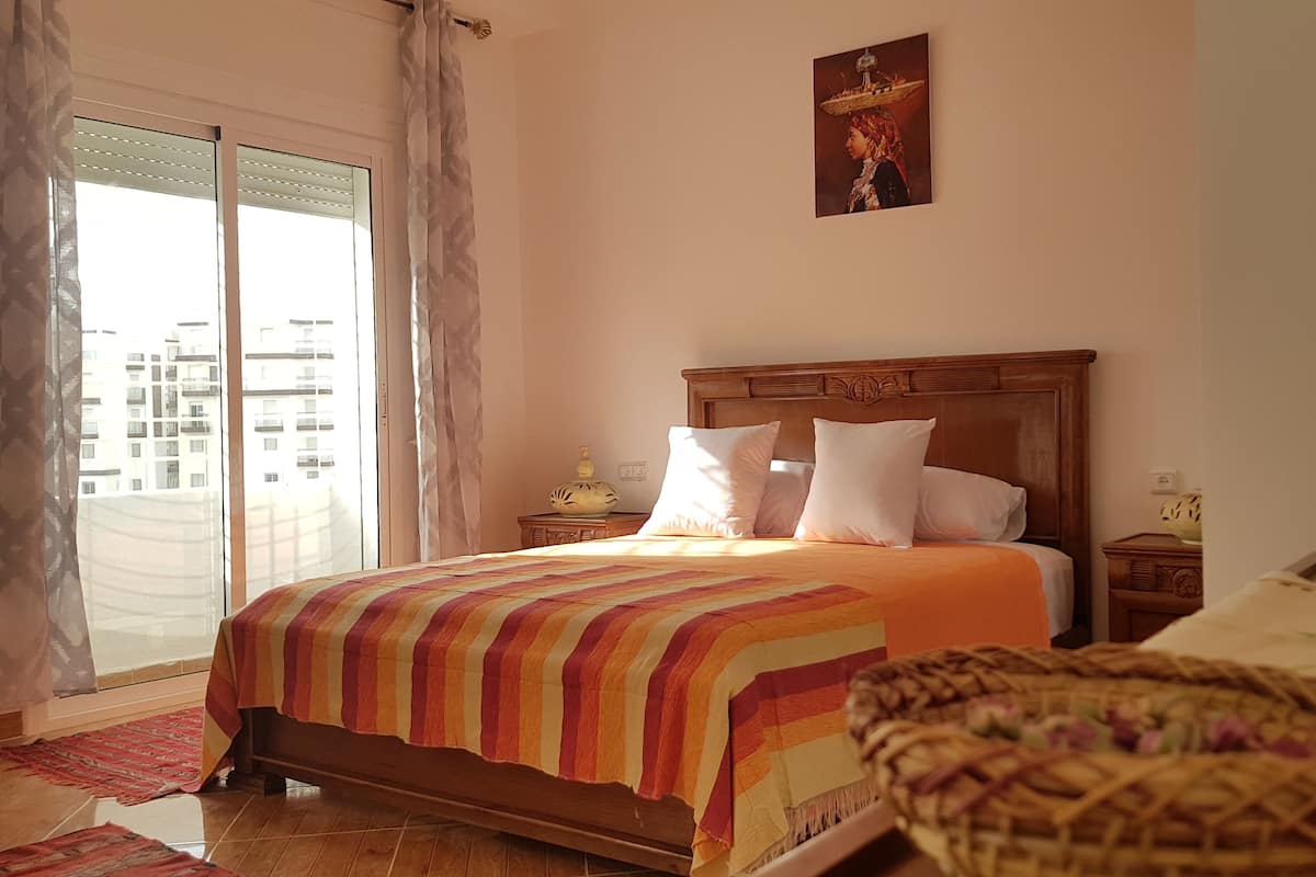 Une chambre avec un grand lit, une table de chevet, une lampe, une fenêtre avec des rideaux et une vue sur les bâtiments à l’extérieur.