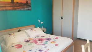 A bed with floral bedding, a nightstand with a lamp, and a painting of a sunset over the ocean on the wall. - La casa del viaggiatore (Catania)