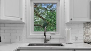 Cocina con gabinetes blancos, pileta de acero inoxidable, ventana con vista a árboles y salpicadero de azulejos.
