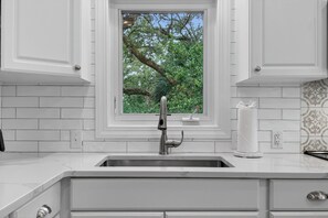 Cocina con gabinetes blancos, pileta de acero inoxidable, ventana con vista a árboles y salpicadero de azulejos.