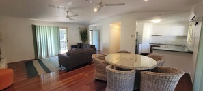 A modern living room with a sofa, a round table with wicker chairs, and a kitchen area with cabinets. - Cabbage Palm Holiday Home (Agnes Water)