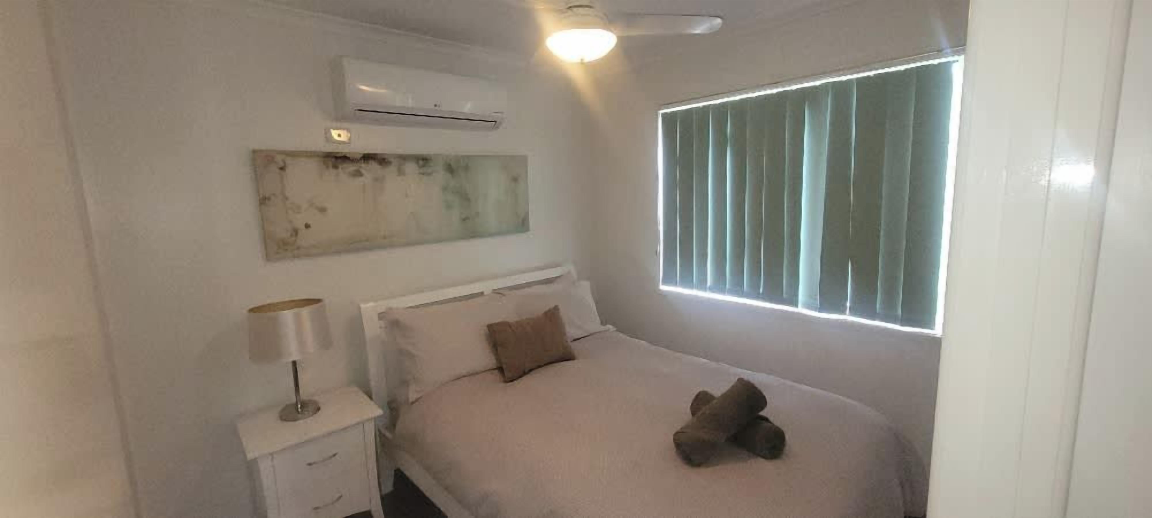 A bedroom with a bed, a nightstand, an air conditioner, a window with blinds, and a ceiling fan.