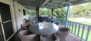 A covered outdoor area with a round table surrounded by wicker chairs, a grill, and a view of a garden with palm trees.