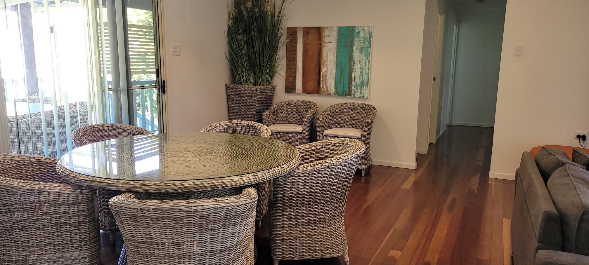 A dining area with a round glass table surrounded by wicker chairs, a potted plant, and a wall with a colorful abstract painting.