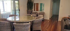 A dining area with a round glass table surrounded by wicker chairs, a potted plant, and a wall with a colorful abstract painting.