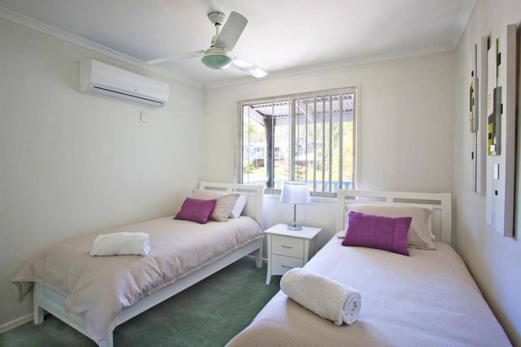 Een slaapkamer met twee bedden, een plafondventilator, een airconditioningapparaat, een raam met tralies en een nachtkastje met een lamp.