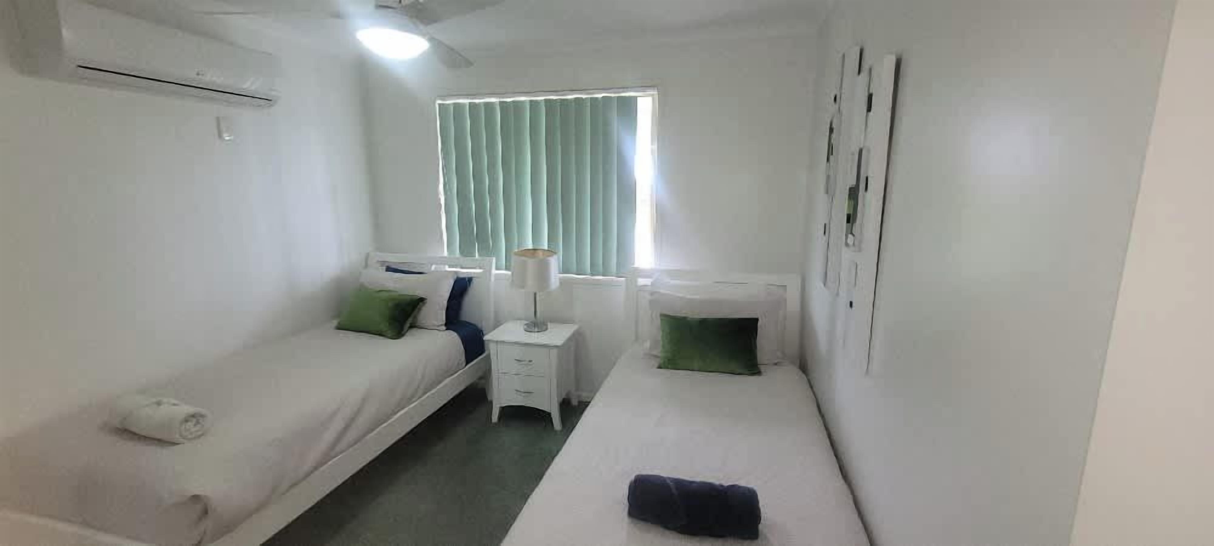 A small, clean room with two single beds, a bedside table with a lamp, and a window with curtains.