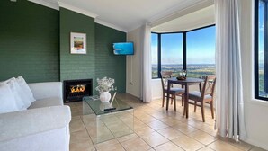 A modern living room with a green accent wall, a fireplace, a glass coffee table, a white sofa, and a dining area with a view of the landscape. - Villa 6 @ Bimbadeen Estate - Spa studio for 2 - Amazing views at every turn (Mount View)