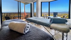A room with two massage tables, a small table with a bottle, and a view of the landscape through large windows. - Villa 6 @ Bimbadeen Estate - Spa studio for 2 - Amazing views at every turn (Mount View)