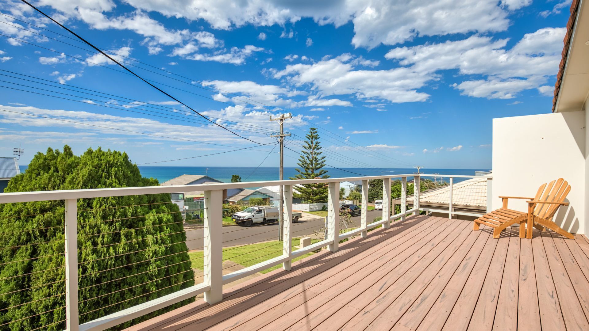 A wooden deck with a white railing, overlooking a residential area and the ocean.