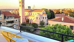 A terrace with two wicker chairs, a view of a historic building with a tower, and a distant crane.