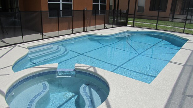 Une piscine avec un espace de jacuzzi circulaire, entourée d’une clôture et, au fond, un bâtiment doté d’un toit recouvert de tuiles.