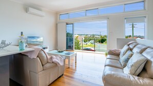 A modern living room with a sofa, a coffee table, and a TV. There is a large window with a view of a residential area.
