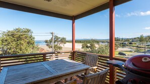 A covered outdoor area with a wooden table and chairs, a grill, and a view of the beach and mountains.