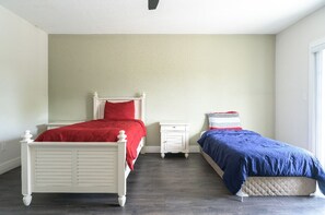 Two single beds with red and blue bedspreads sit against a light-colored wall, accompanied by a small nightstand with drawers.