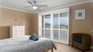 A bedroom with a bed, a dresser, a chair, a ceiling fan, and a large window with blinds. - Mistral, Gerroa, South Coast (Gerroa)