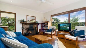 A living room with a fireplace, a television, and a large window with a view of trees. - Charming Cottage Retreat by HolidayCo. (KILLCARE)