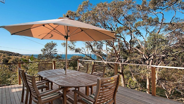 A wooden deck with a round table and four chairs, an umbrella, and a view of the sea and trees. - Charming Cottage Retreat by HolidayCo. (KILLCARE)
