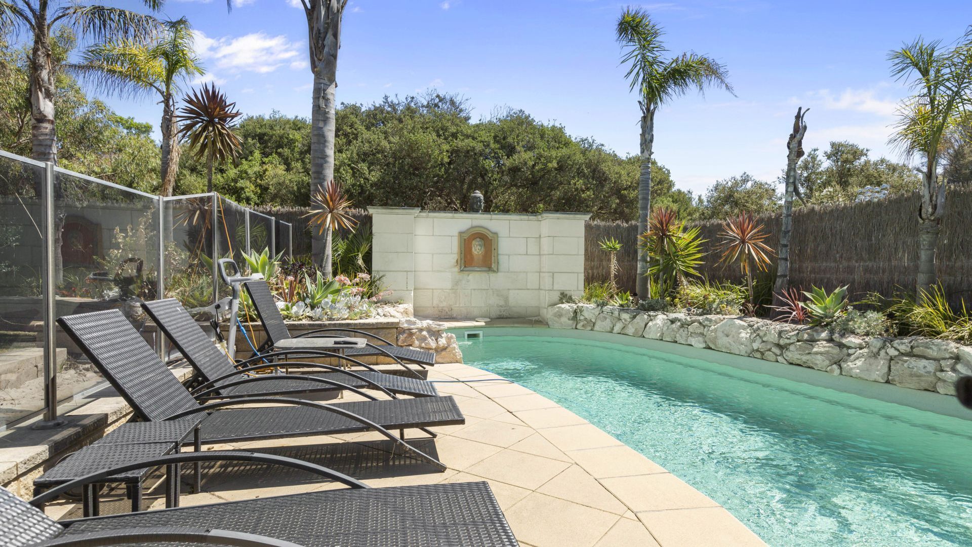 A pool area with lounge chairs, a stone wall, and tropical plants.