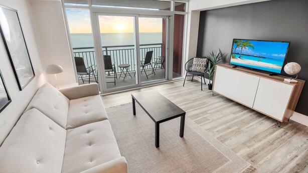 A modern living room with a sofa, a coffee table, a TV on a cabinet, and a balcony with a view of the sea.
