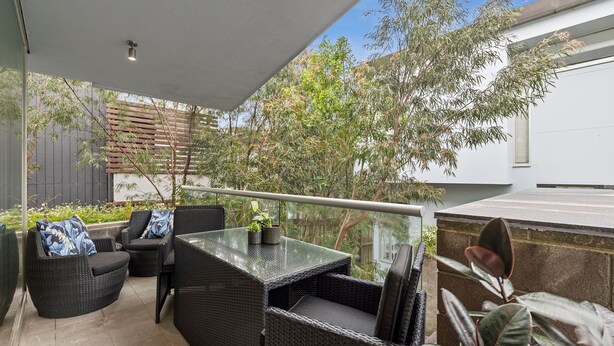 A covered outdoor seating area with wicker furniture and a glass table, surrounded by greenery and a view of a building. - A Touch Of Class (Lorne)
