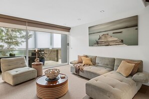 A modern living room with a grey sofa, armchairs, and a coffee table. There is a large window with a view of trees and a building outside. - A Touch Of Class (Lorne)