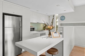 A modern kitchen with stainless steel appliances, a white countertop, and a bar area with stools. - A Touch Of Class (Lorne)