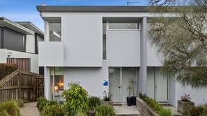 A modern two-story residential building with a flat roof, large windows, and a small front garden with plants and a wooden fence. - A Touch Of Class (Lorne)