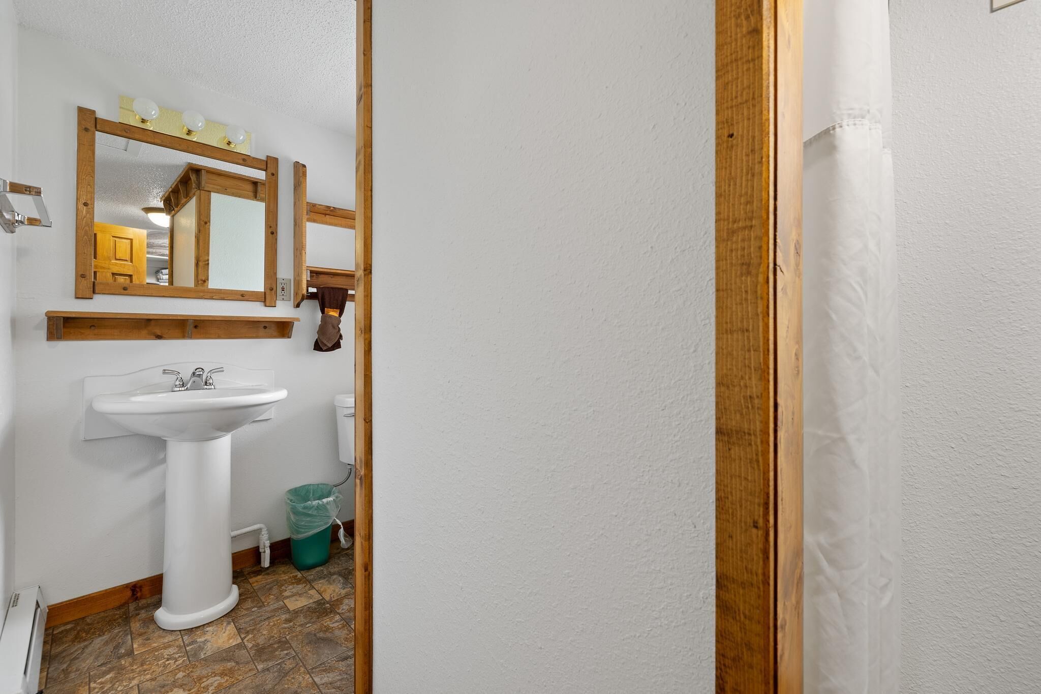 Un baño con un lavamanos blanco de pedestal, un espejo con marco de madera y un cubo de basura verde.