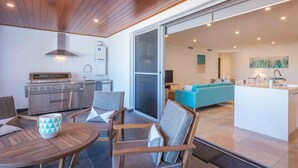 A modern kitchen with a built-in oven and a water dispenser, a dining area with wooden furniture, and a living space with a sofa and a TV. - Blue Waves, Kiama, South Coast (Kiama)
