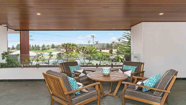 A covered outdoor seating area with four wooden chairs and a round table, overlooking a residential area with houses and greenery. - Blue Waves, Kiama, South Coast (Kiama)