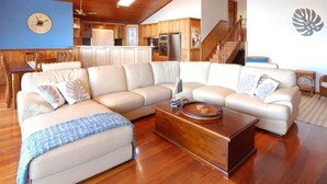 Un salón espacioso con un amplio sofá modular en forma de L, suelo de madera, una mesa de centro de madera y una cocina al fondo.