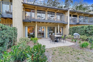 A two-story building with a patio area featuring a table and chairs, surrounded by various plants and a mailbox.