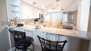 A modern kitchen with white cabinets, a granite countertop, and a built-in microwave. There are two black bar stools and a small table with a chair. - Oceanfront 2BR Condo 3rd Floor Balcony Views, Unit 307 (Indian Harbour Beach)