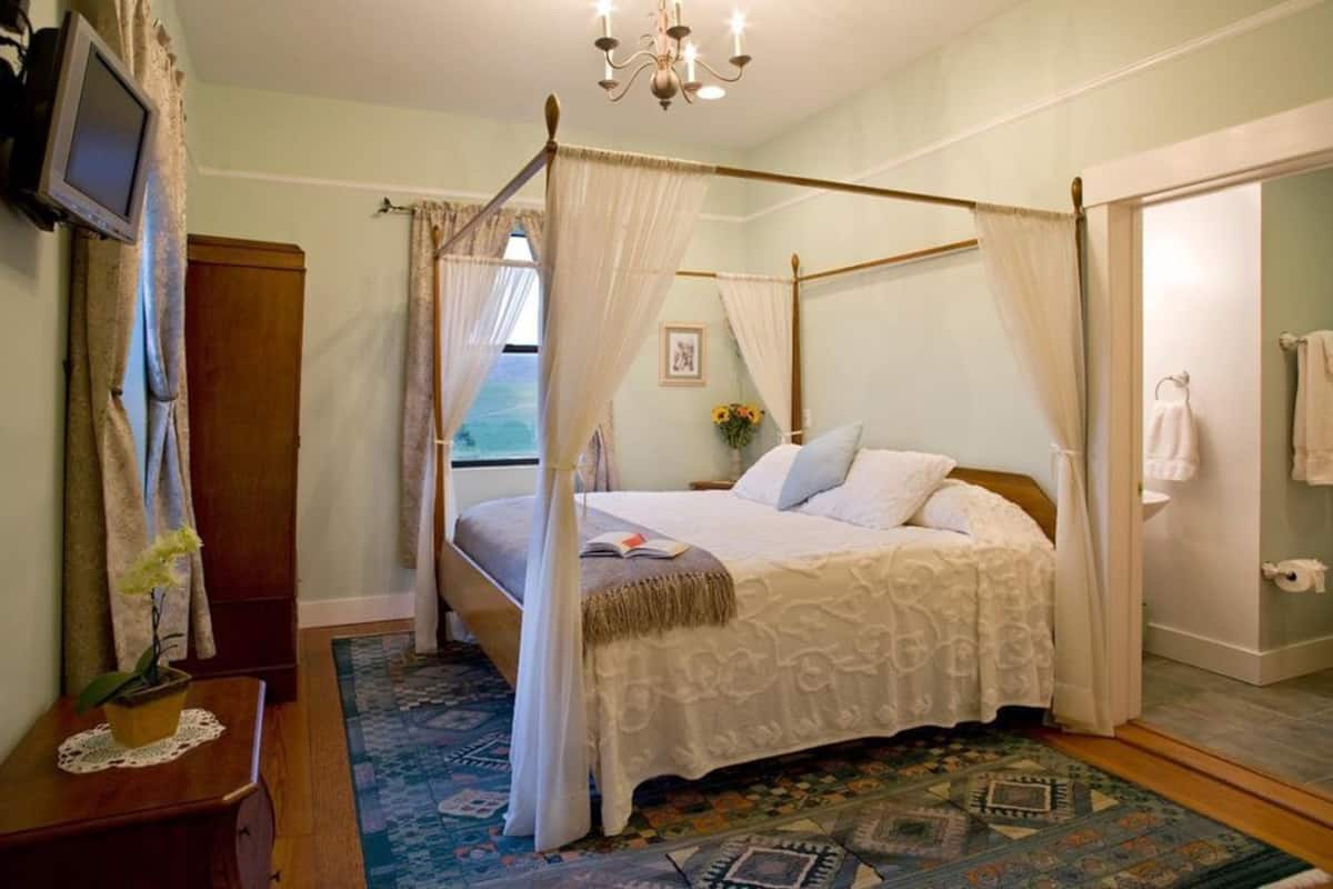 A four-poster bed with a canopy, a wooden nightstand with a plant, a patterned rug, and a view of the sea through the window.