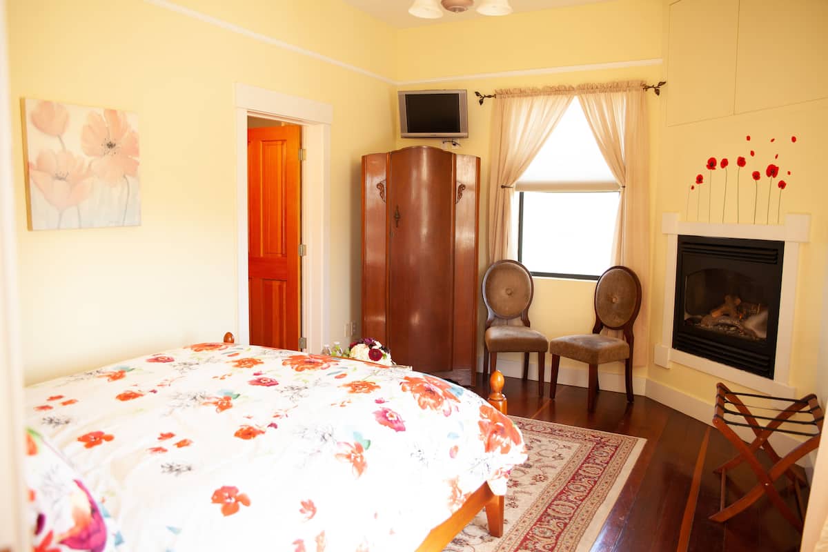 A bedroom with a bed, two chairs, a television, a wardrobe, a fireplace, and a window with curtains.
