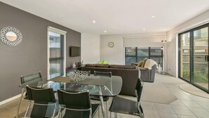 A modern living room with a glass dining table, black chairs, a brown sofa, and a large window.