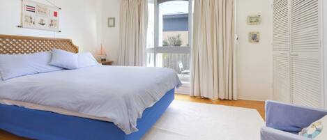 A bedroom with a large bed, a chair, a window with curtains, and a wall with a framed picture.