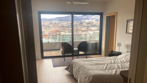 Un dormitorio con un ventanal grande que ofrece vistas a la ciudad, una cama, un sillón y una mesita.