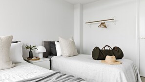 A minimalist bedroom with two beds, a bedside table with a lamp, a hanging clothes rack, and a large bag on the floor.